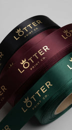 Three rolls of satin ribbon in black, burgundy, and forest green, each cutom  printed with the gold 'LOTTER PRINT CO.' logo, arranged diagonally on a light gray background.