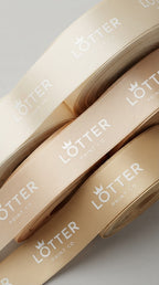A close-up of several rolled ribbons in warm cream, beige, and tan tones, each custom printed and branded with the white Lotter Print Co. logo and name.
