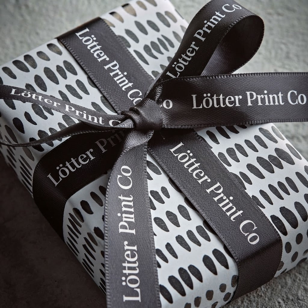 A gift box wrapped in white paper with a custom black abstract dot pattern and tied with a wide custom  black ribbon printed with the brand name 'Lötter Print Co'.