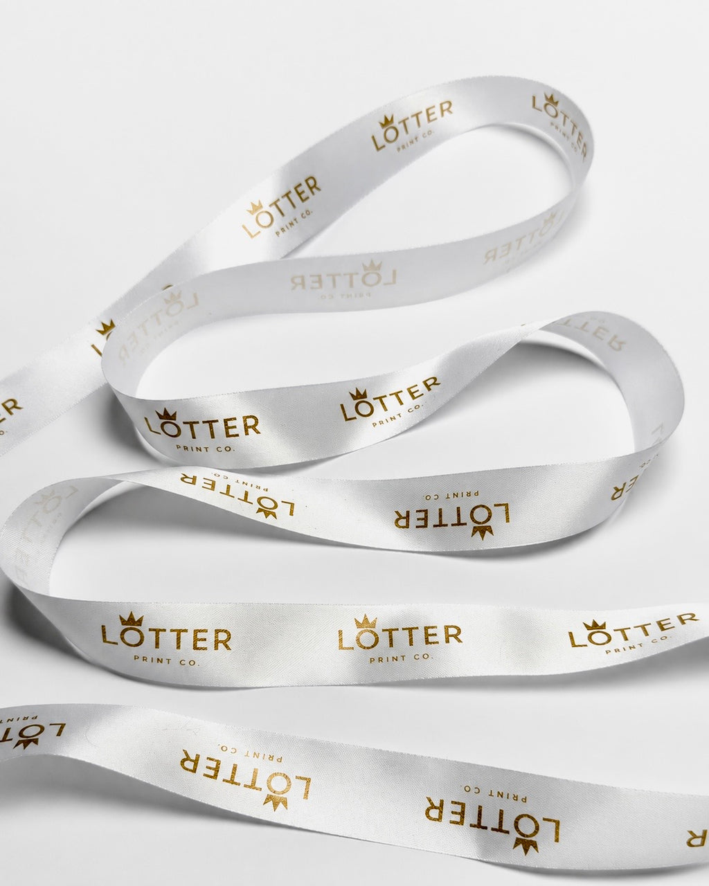White satin ribbon with gold foil 'LOTTER PRINT CO.' branding printed repeatedly, arranged in loops on a light gray background.