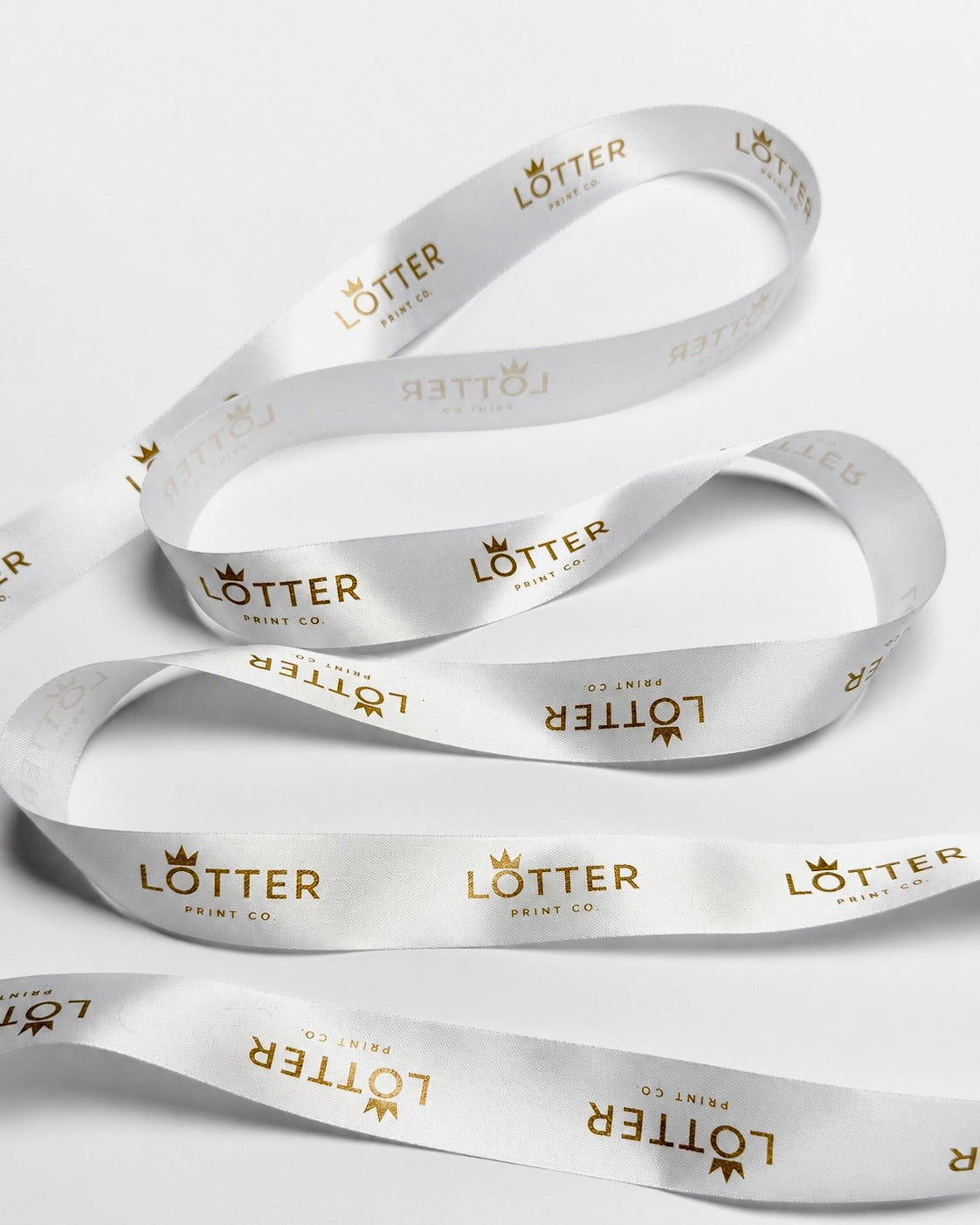 White satin ribbon with gold foil 'LOTTER PRINT CO.' branding printed repeatedly, arranged in loops on a light gray background.