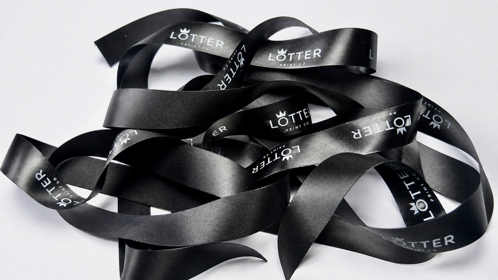 A pile of black satin ribbon with white 'LOTTER' branding printed repeatedly on it, photographed on a plain white background.
