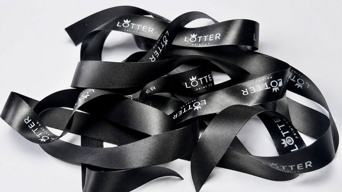 A pile of black satin ribbon with white 'LOTTER' branding printed repeatedly on it, photographed on a plain white background.