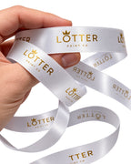 A hand holding a white satin ribbon printed with the gold 'LOTTER PRINT CO' logo, shown against a clean white background.